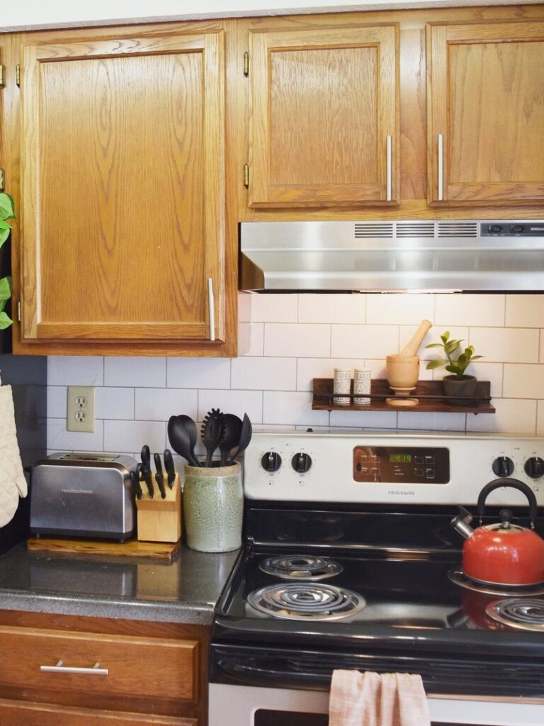Oak cabinets, subway-look backsplash, dark wood floating shelf and red kettle on stove