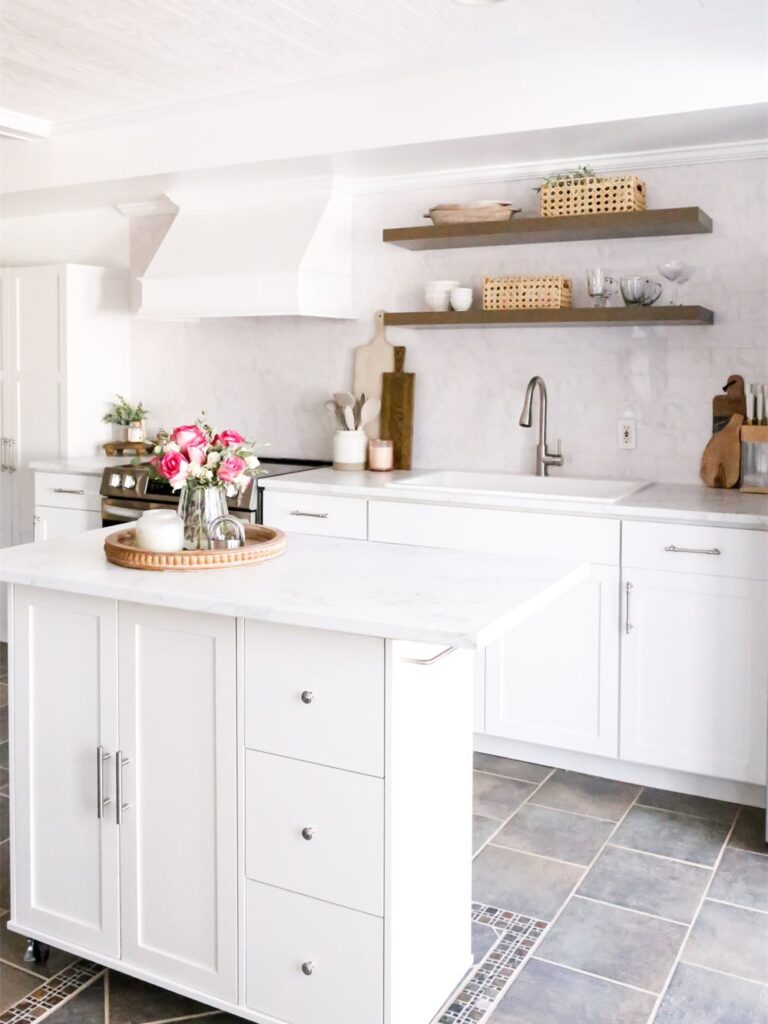 White kitchen with rolling island, white range hood, dark floating shelves and slate floor